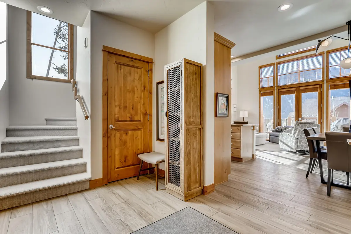 Entryway with wooden door, stairway with light carpet, bench, and mesh cabinet. Open living area with large windows, mountain views, wooden floors, dining table, and sofa in the background. Bright and modern interior.