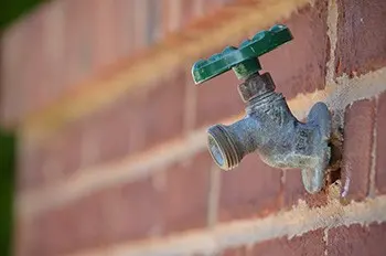 Outdoor metal water faucet with a green handle attached to a red brick wall, photographed in focus with a blurred background.