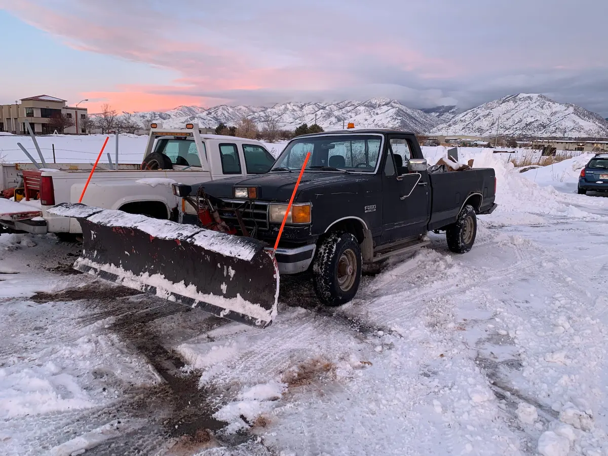A black pickup truck with a snow plow attached is parked in a snowy lot near another truck. Snow-covered mountains and buildings are visible in the background during sunset.