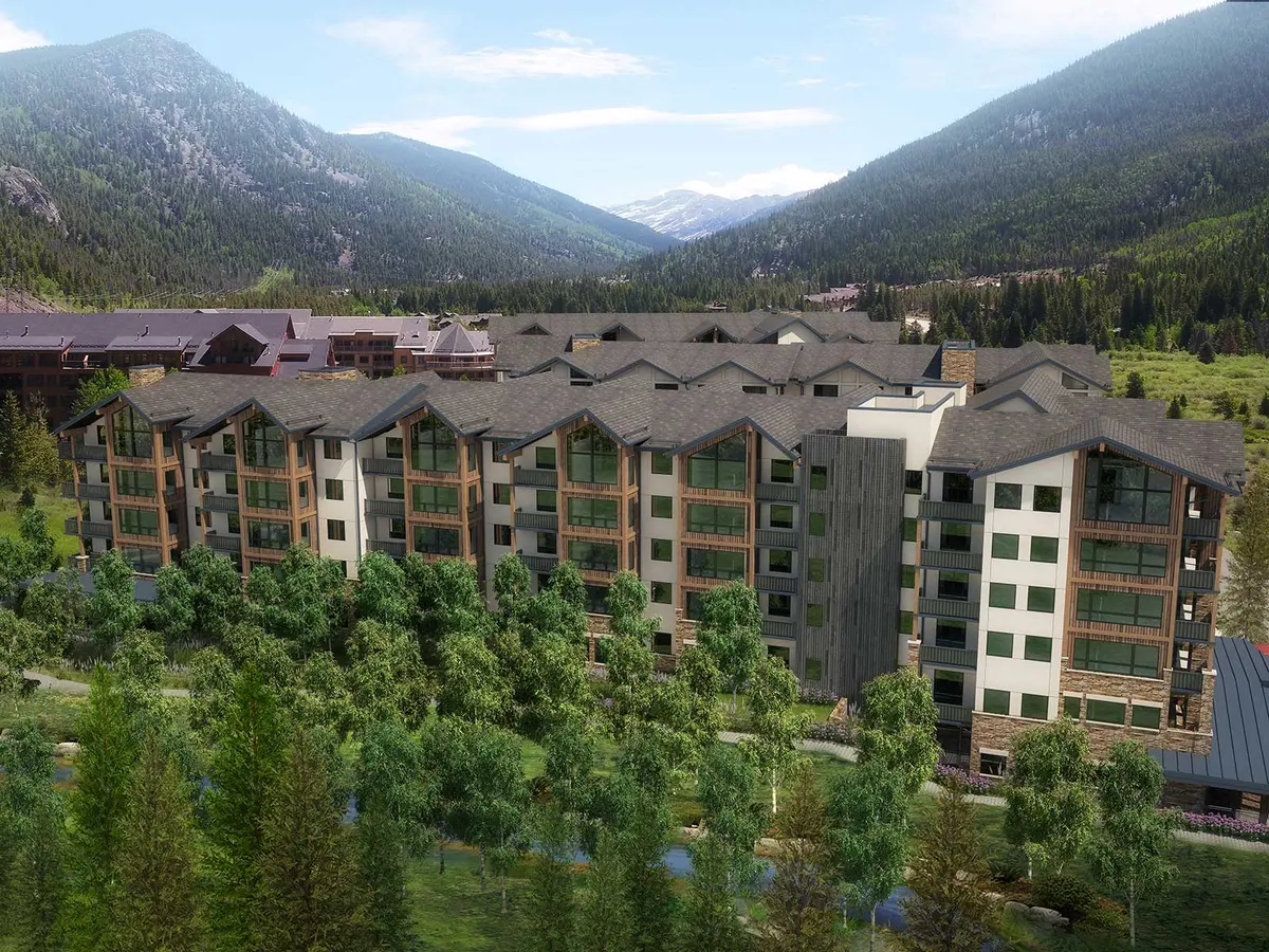 A large, multi-story mountain resort building with balconies and large windows, surrounded by green trees and mountains under a partly cloudy sky.