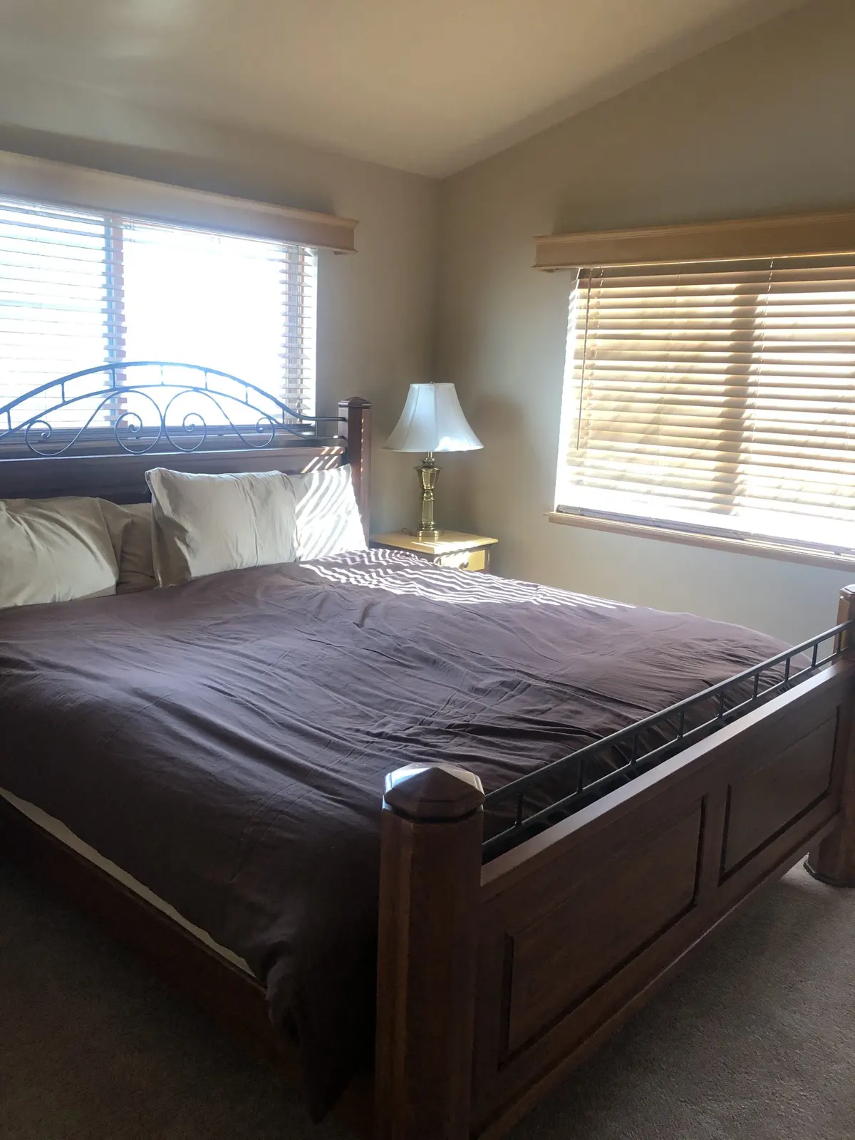 A sunlit bedroom with a large wooden bed, brown bedding, two pillows, a side table with a lamp, and windows with closed blinds letting in light.
