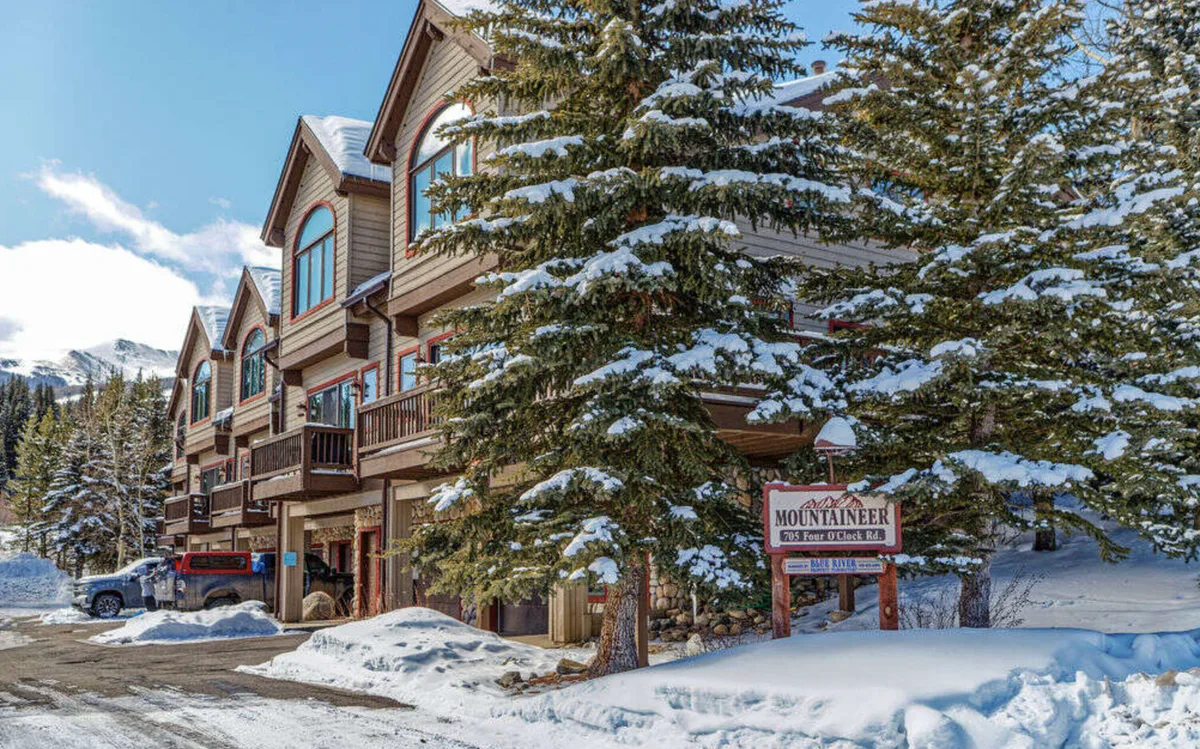 Snow-covered townhouses with balconies and garages sit among evergreen trees. A sign reads Mountaineer and mountains are visible in the background under a bright blue sky.