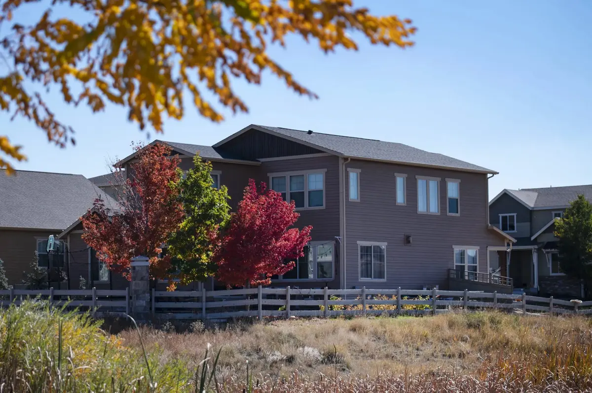 Two-story suburban house with brown siding, surrounded by autumn trees with red and yellow leaves, a white fence, and dry grass in the foreground under a clear blue sky.