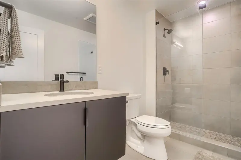 Modern bathroom with a gray vanity, round sink, black fixtures, white toilet, and a glass-enclosed shower with gray tile walls and a built-in shelf. Towels hang on a rack near the door.