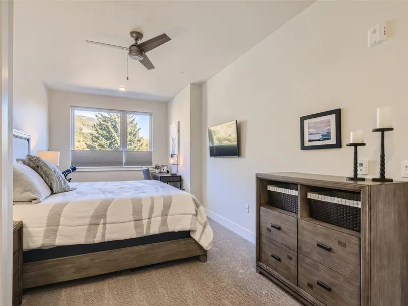 Modern bedroom with a large bed, wooden dresser, desk by the window, TV on the wall, neutral decor, carpeted floor, and a ceiling fan. Sunlight streams in through a big window showing trees outside.