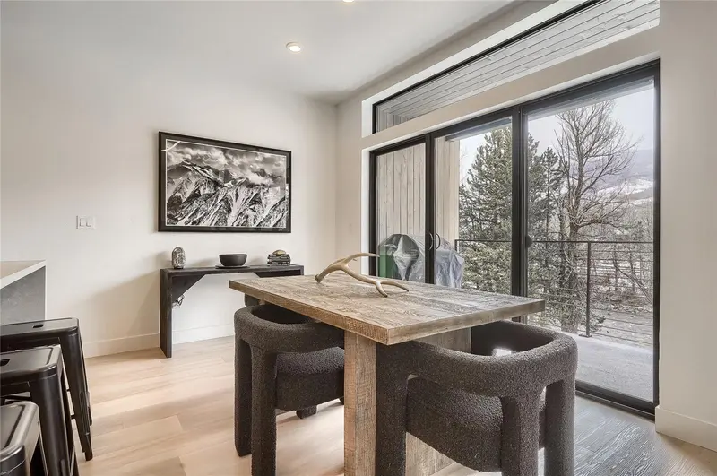 Modern dining area with a rustic wooden table, black chairs, decorative antlers centerpiece, large glass doors leading to a balcony, and a black-and-white mountain photo on the wall. Trees and mountains are visible outside.