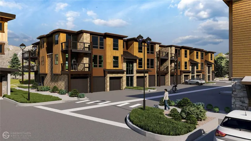 A modern townhouse complex with three-story units, stone and wood exteriors, large windows, and balconies. People walk and cycle on the street, and cars are parked nearby under a partly cloudy sky.