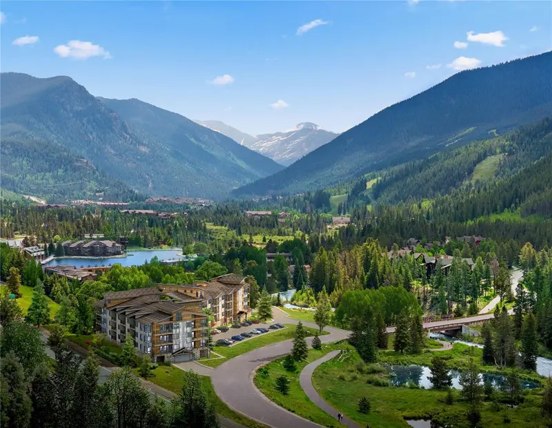 Aerial view of a mountain valley with a resort, parking lot, trees, small lakes, and winding roads surrounded by forested hills under a blue sky.
