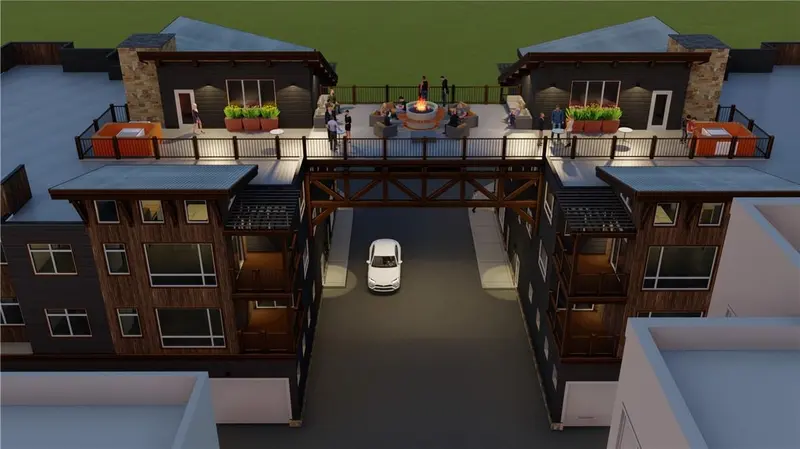 Aerial view of two modern buildings connected by a rooftop patio with people socializing, planter boxes, and a fire pit; a car drives underneath through the passage below.