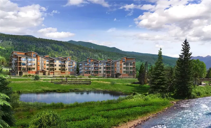 Modern apartment buildings sit near a pond and river, surrounded by lush green grass, evergreen trees, and mountains under a partly cloudy sky.