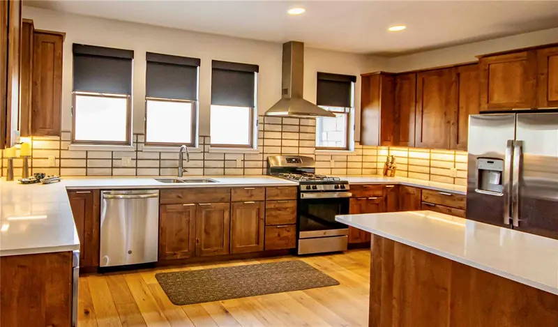 Modern kitchen with wooden cabinets, stainless steel appliances, white countertops, subway tile backsplash, three windows with black shades, and light wood flooring. A woven rug is placed in front of the sink area.