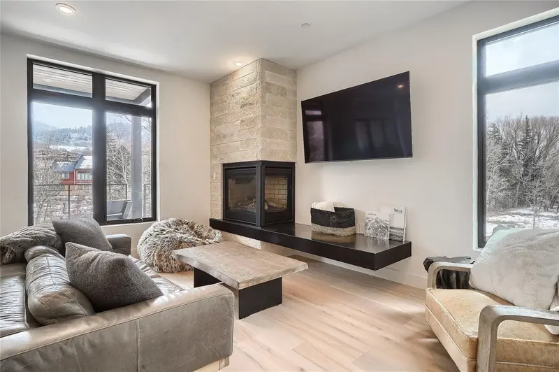 Modern living room with a large TV above a floating shelf, a stone fireplace, cozy sofas with pillows and throws, large windows, and light wood floors. Snowy trees and mountains are visible outside.