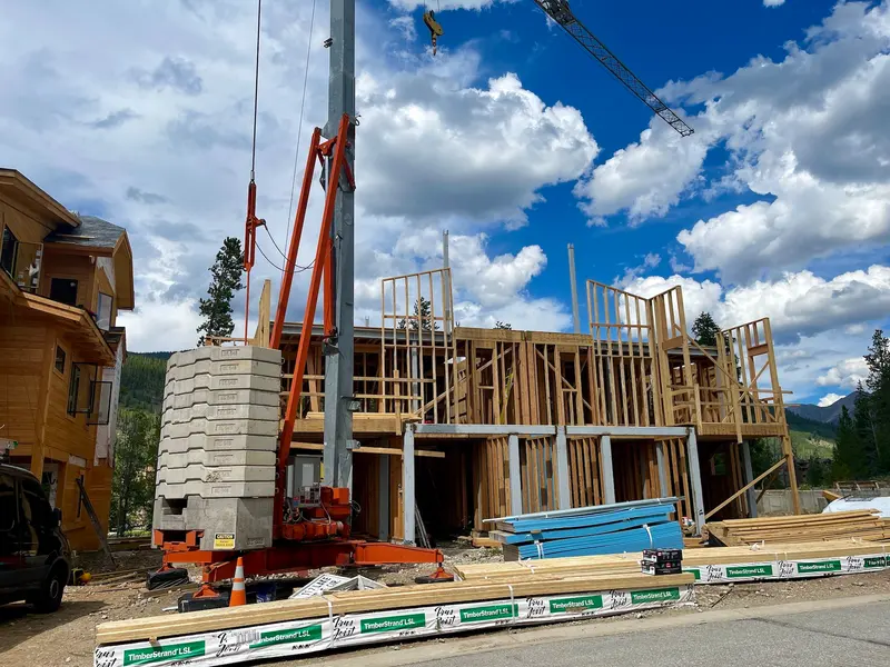 Wood-framed building under construction, with a crane, construction materials, and blue sky with clouds in the background. Scaffolding and partially built walls are visible.