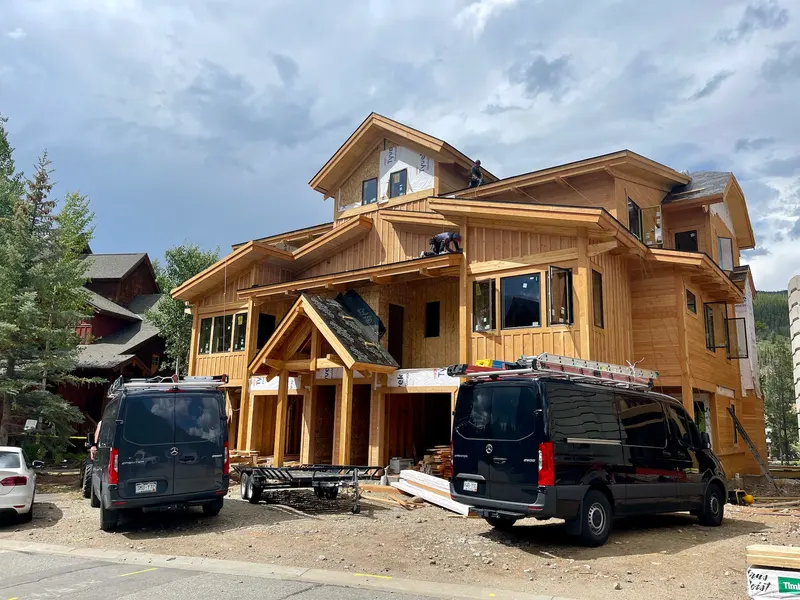 A large wooden house under construction, with two black vans parked in front. A worker is on the roof, and building materials are scattered around. The sky is partly cloudy.