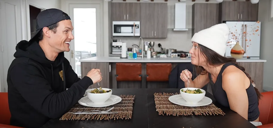 Two people sit across from each other at a dining table, smiling and eating from bowls. A modern kitchen is visible in the background. Both are wearing casual clothes, and the woman is wearing a white beanie.